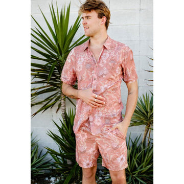 Front view of a man wearing a matching short sleeve shirt and shorts printed with white banksia flowers on a light pink background.