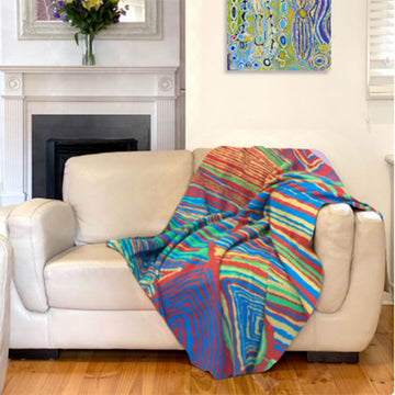 A cotton blanket printed with colourful artwork by Judy Watson is laying over a light coloured couch with a fireplace and painting in the background.