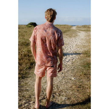 Front view of a man wearing a matching short sleeve shirt and shorts printed with white banksia flowers on a light pink background.