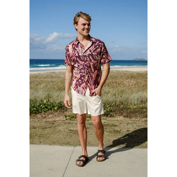 Front view of a man wearing a burgundy short sleeve shirt printed with  flowers with cream shorts standing in front of a beach.