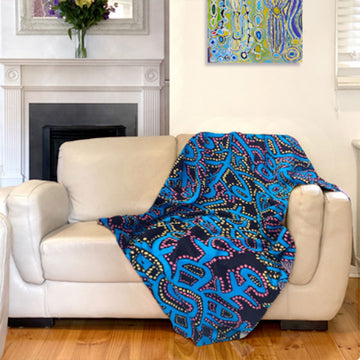 A blue throw printed with artwork by Theo Hudson is laying on a light coloured couch with a painting and fireplace in the background.