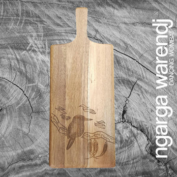 Wooden cutting board with engraved kookaburra design on a gray wooden surface, featuring the brand 'ngaga warendj'.