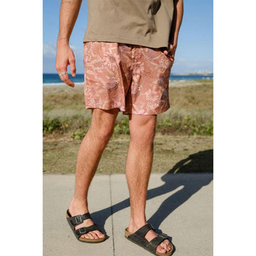 Close up front view of a man wearing an olive green tshirt with floral printed rust coloured shorts in front of a beach.