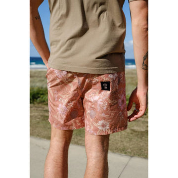 Close up front view of a man wearing an olive green tshirt with floral printed rust coloured shorts in front of a beach.