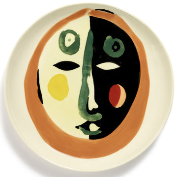 A ceramic plate  painted with an abstract face in orange and green.
