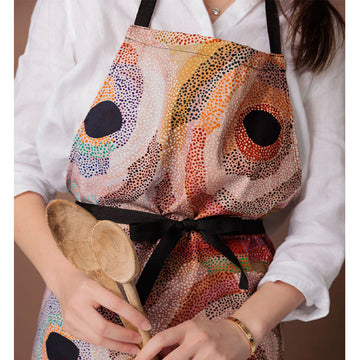 A cotton apron is lying flat and printed with artwork by Marianne Burton with black straps.