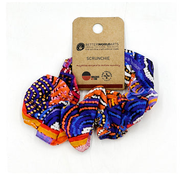 A scrunchie printed with artwork by Nora Davidson in blue and orange with a brown product label attached.