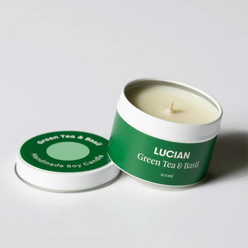 A tin with a candle inside resting on the lid. The tin and lid have the text 'Lucian Green Tea and Basil'.