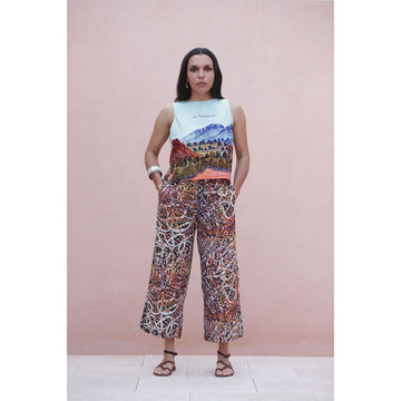 Woman wearing a sleeveless top with a colorful landscape design and patterned pants against a pink background.