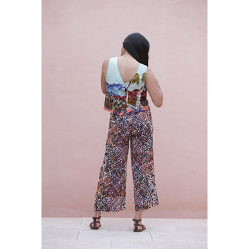 Woman wearing a sleeveless top with a colorful landscape design and patterned pants against a pink background.