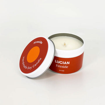 A candle inside a tin with the lid resting on the side. The tin and lid have a red label with the text 'Lucian Fireside'.