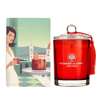 A red glass candle next to its box. The box has an illustration of a woman holding a drink on a balcony looking over a city.