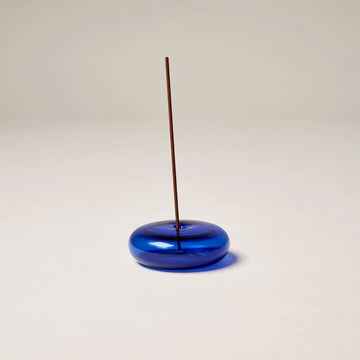 A round glass incense holder in blue with an incense stick in the middle.