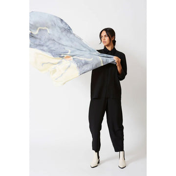 A woman is holding out a silk scarf printed with artwork in light grey blue and light yellow.