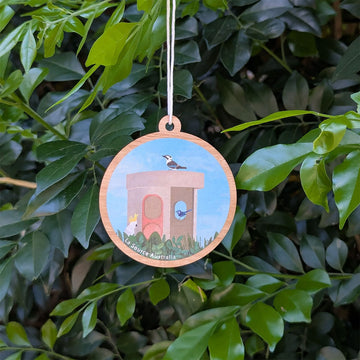 Decorative hanging ornament with an illustration of a Canberra bus stop and birds, hanging against a green leafy background.