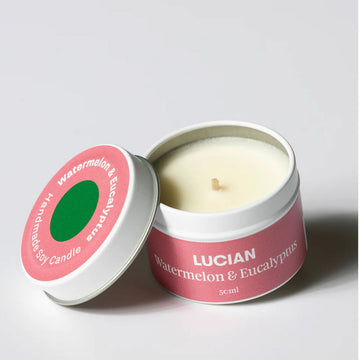 A candle inside a small tin with the lid leaning on the side. The lid and tin have a pink label with the text 'Lucian Watermelon& Eucalyptus'.