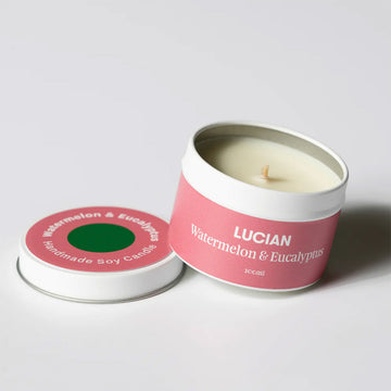 A candle in a tin resting on the lid. The lid and tin have a pink label with the text 'Lucian Watermelon & Eucalyptus'.
