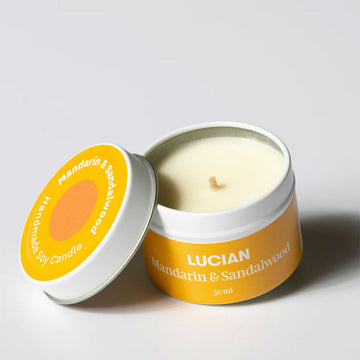 A candle in a tin with the lid resting next to it.  The tin and lid have an orange label with the text 'Lucian Mandarin & Sandalwood'.