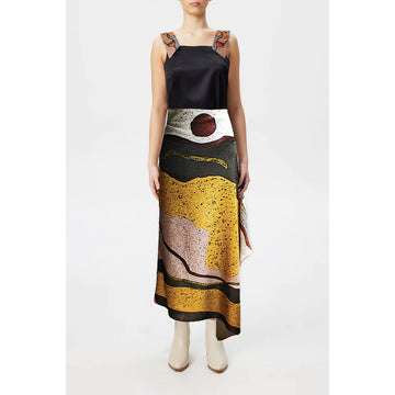 A close up of a model wearing a black tank top with patterned straps and a large silk scarf wrapped around her waist like a skirt.