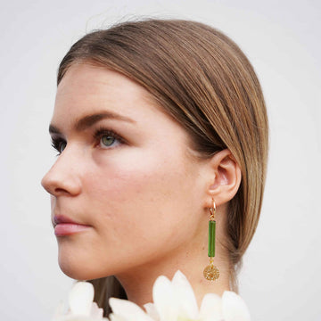 A pair of gold hoop earrings with a long green bead and gold glitter circle attached to the hoop.