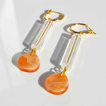 A pair of gold hoop earrings with a long clear bead and orange marble circle attached to the hoop.