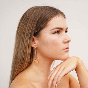 A pair of gold hoop earrings with a long clear bead and orange marble circle attached to the hoop.