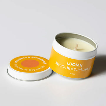 A candle inside a small tin resting on the lid. The tin and lid have an orange label with the text 'Lucian Mandarin & Sandalwood'.