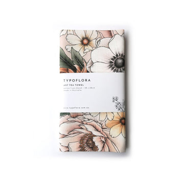 A folded up tea towel in beige illustrated with large flowers in white, light pink and yellow. There is a white sleeve around the tea towel with product information.