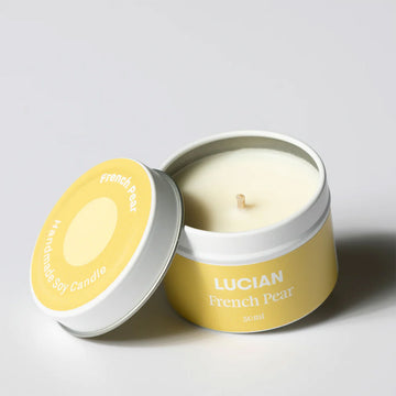 A candle inside a tin with the lid leaning on the side. The lid and tin have a light yellow label with the text 'Lucian French Pear'.