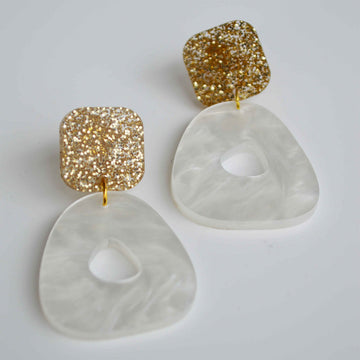 A pair of drop earrings with a gold glitter square on top and a pearl white circle on the bottom.