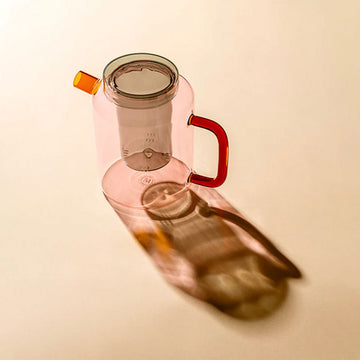 Pink glass pot with a red handle on a beige background.