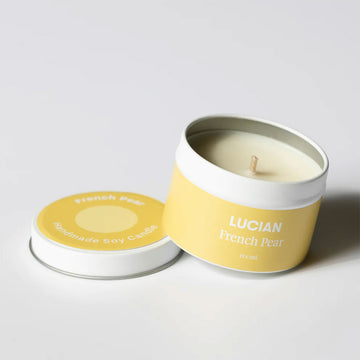 A candle in a in leaning on the lid. The tin and lid have a light yellow label with the text 'Lucian French Pear'.