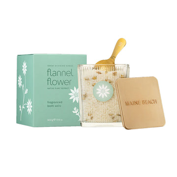 A green box with the heading 'flannel flower' in dark green is behind a clear glass filled with bath salts with a gold scoop inside.