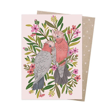 Greeting card with two galahs on a branch surrounded by flowers and leaves.