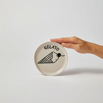 A hand is holding up a round white dish with a black line drawing of an ice cream cone and the text 'Gelato'.