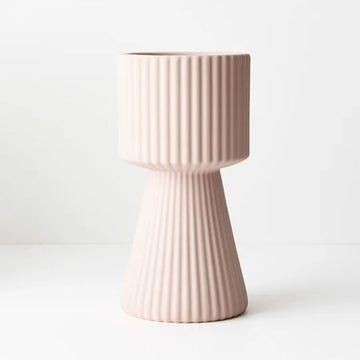 A tall light pink ceramic vase with a round shape on the top half and a triangular base embossed with vertical lines.