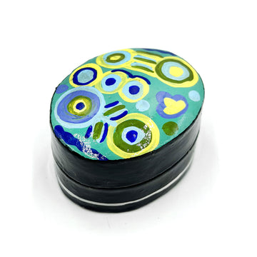 A black pill box printed with artwork by Cedric Varcoe in green and blue on the top.