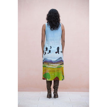 Woman wearing a sleeveless dress with a colorful landscape design with birds flying against a pink background.