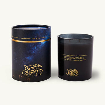 Black candle with gold text next to its packaging on a white background.