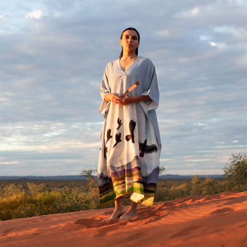 Woman in a long, patterned kaftan standing on a red dirt landscape with a cloudy sky.