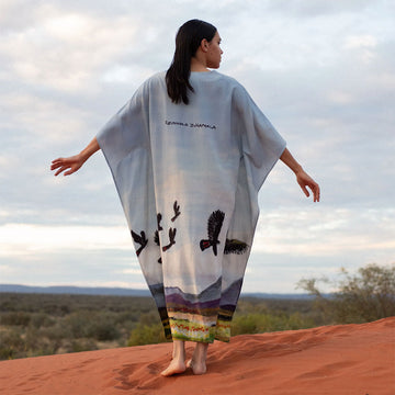 Woman in a long, patterned kaftan standing on a red dirt landscape with a cloudy sky.