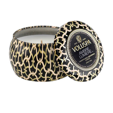 A tin candle printed with a black and gold pattern with the text 'Volupsa Ambre Lumiere' on the lid.