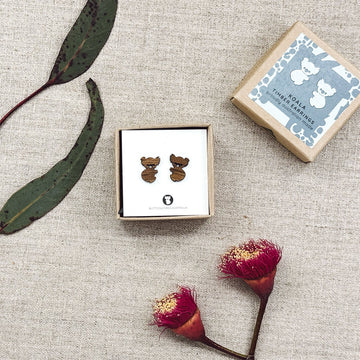 Wooden koala-shaped earrings in a box on a textured surface with leaves and flowers.