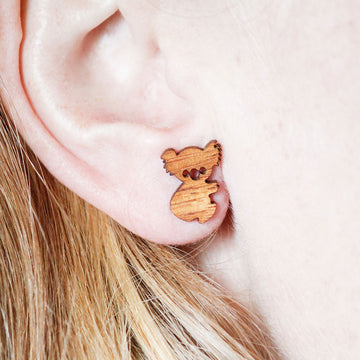 Wooden koala-shaped earrings in a box on a textured surface with leaves and flowers.
