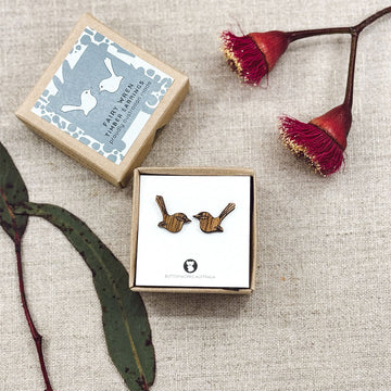 A pair of wooden fairy wren earrings in a box surrounded by leaves and flowers.
