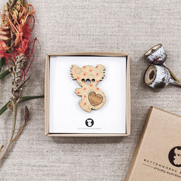 Wooden koala shaped brooch in a box on a textured surface with flowers and packaging.