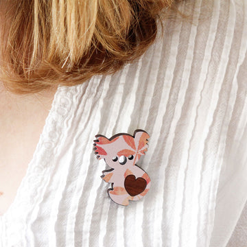Wooden koala shaped brooch in a box on a textured surface with flowers and packaging.