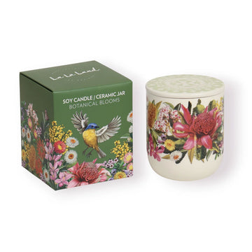 A candle in a ceramic jar next to a green box titled 'Soy Candle/Ceramic Jar Botanical Blooms' with illustrations of flowers and a bird.