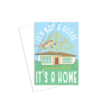 A greeting card with an illustration of a house with a large gate in front and a plane flying above with the text 'It's Not  a House Its a Home'.