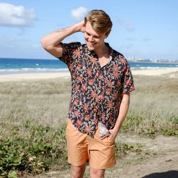 A man is wearing a floral shirt and orange shorts with a beach in the background.
